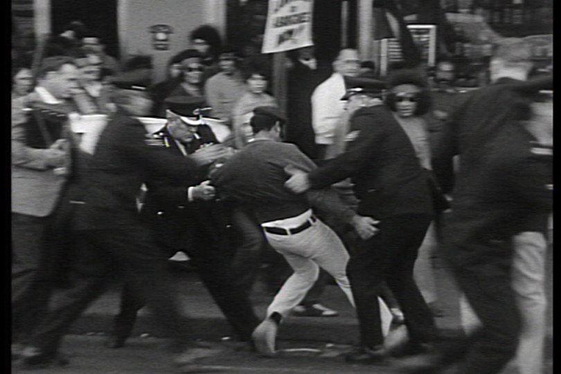 A black and white photo showing an Aboriginal man reeling back on his heels after being thrown by three police officers.