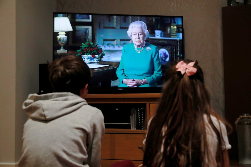 Two children watch the Queen on television, their backs to the camera.