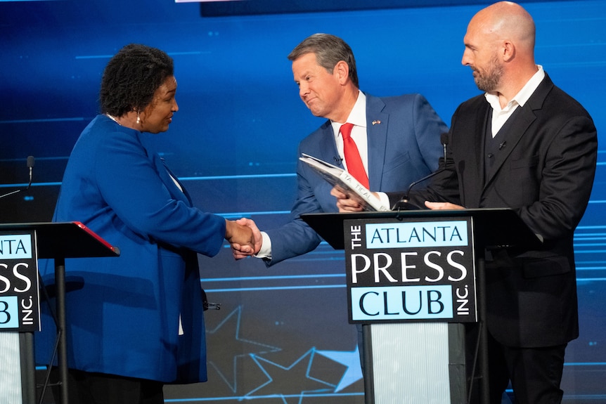 Stacey Abrams shakes hands with Governor Brian Kemp on stage at the Atlanta Press Club, both are wearing blue suits