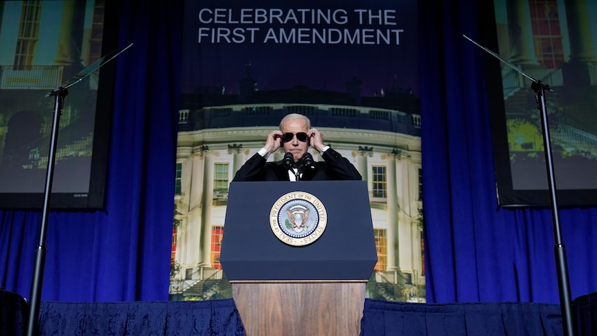 Joe Biden stands at a podium on stage putting on a pair of dark sunglasses. 