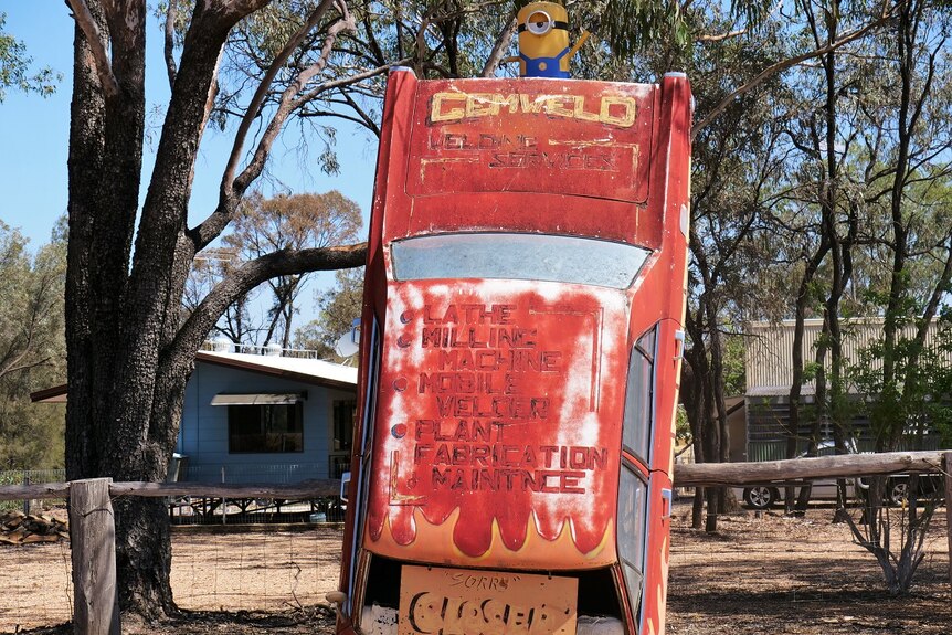 Old red car stands against a fence with a minion statue on top, Gemweld and other text written on car.