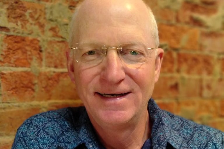 A man with a blue shirt and a bald head smiles with a brick wall behind him