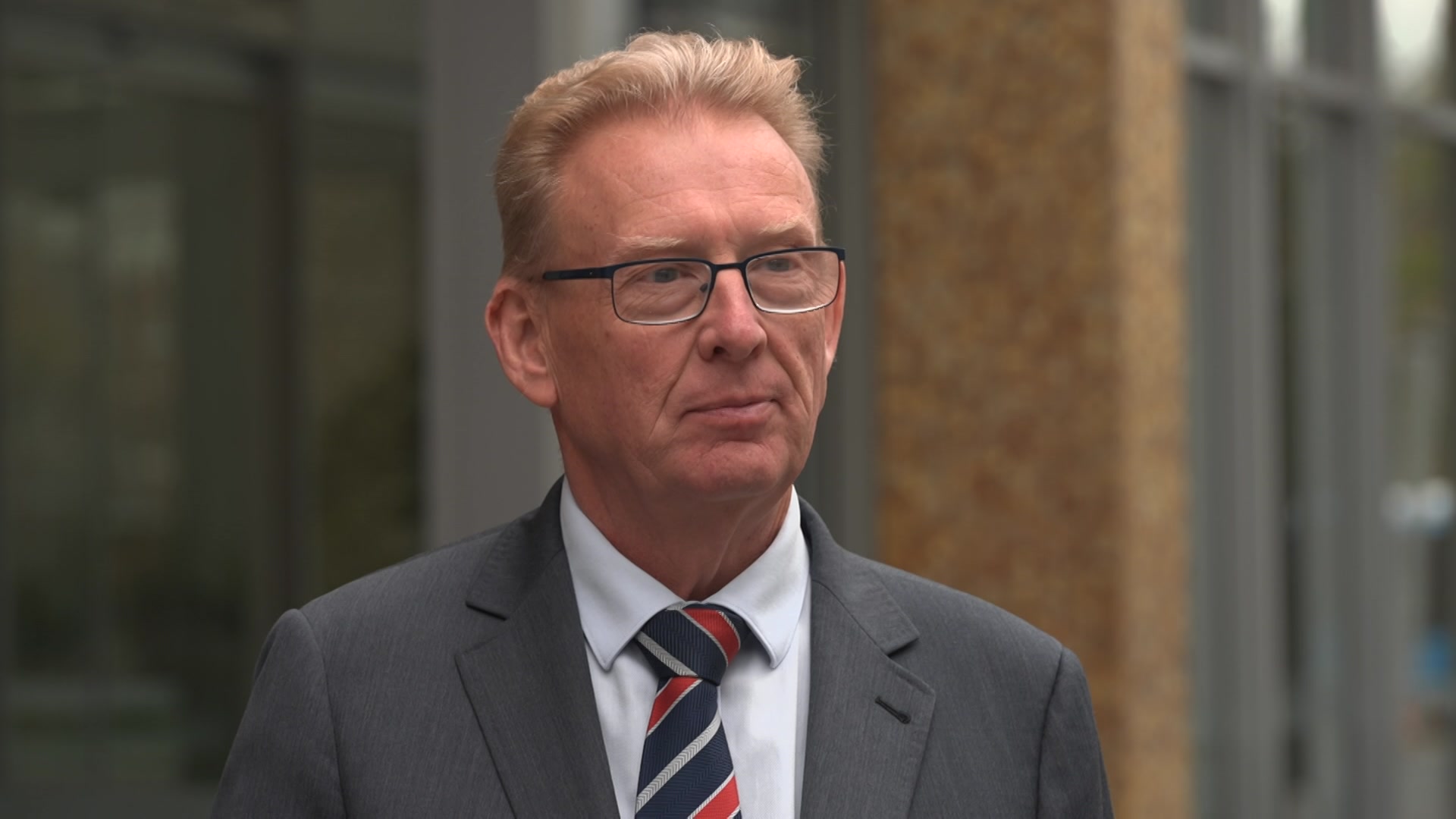 A man with short blonde hair wearing a suit and tie stands looking serious outside a building with gold-tiled columns. 