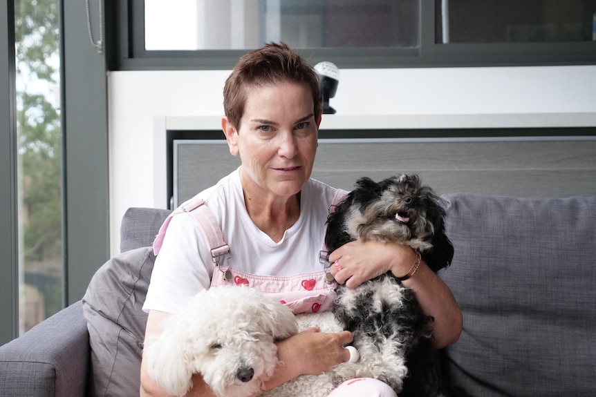 A woman wearing pink overalls over a white t-shirt smiles, with two dogs in her lap