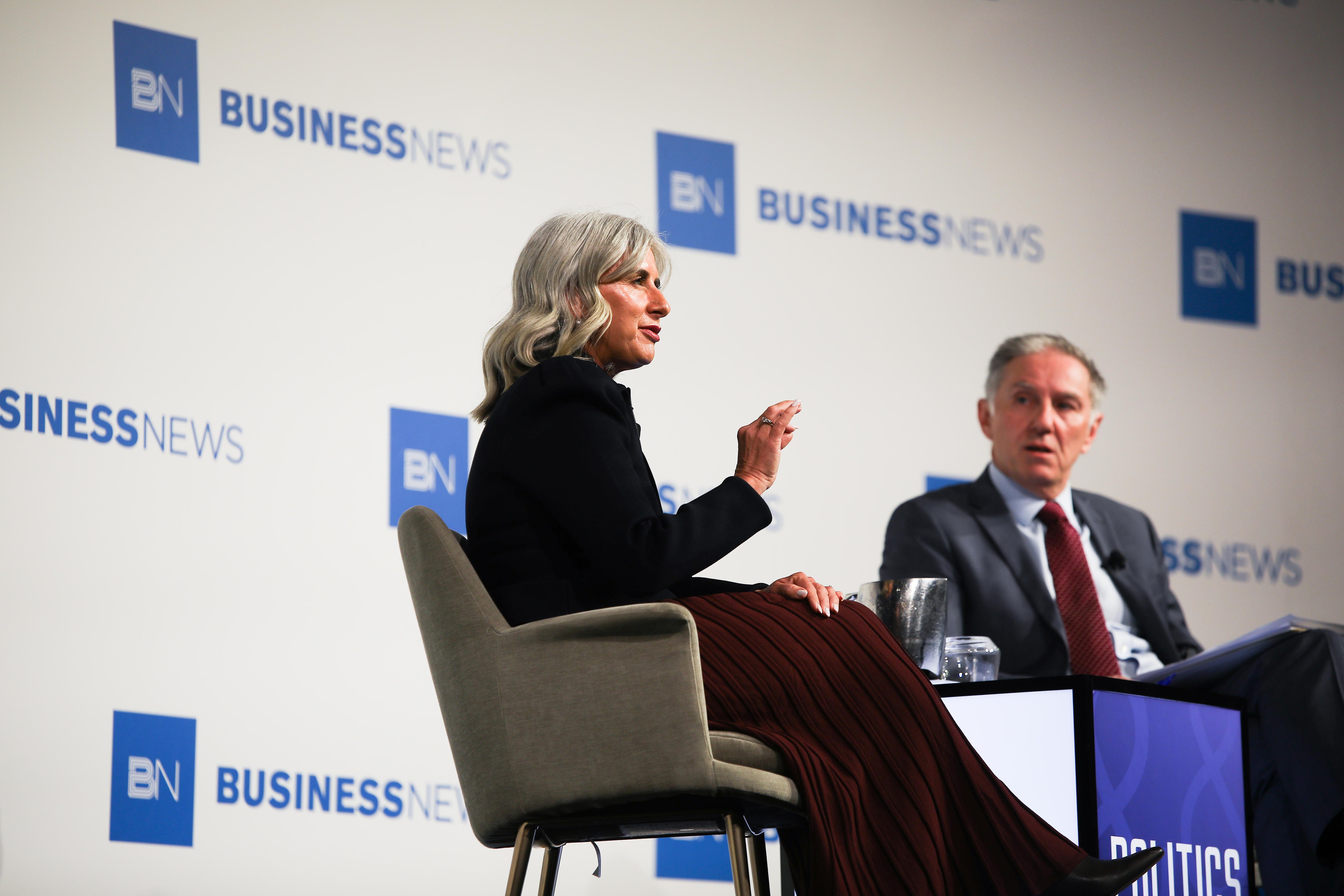 A woman and man sit on chairs in front of a large Business News poster