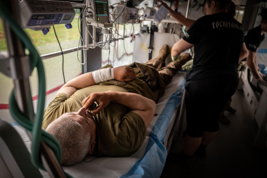 A man lays in a hospital bed inside a bus, while a medic wearing surgical gloves leans in to adjust a monitor above him