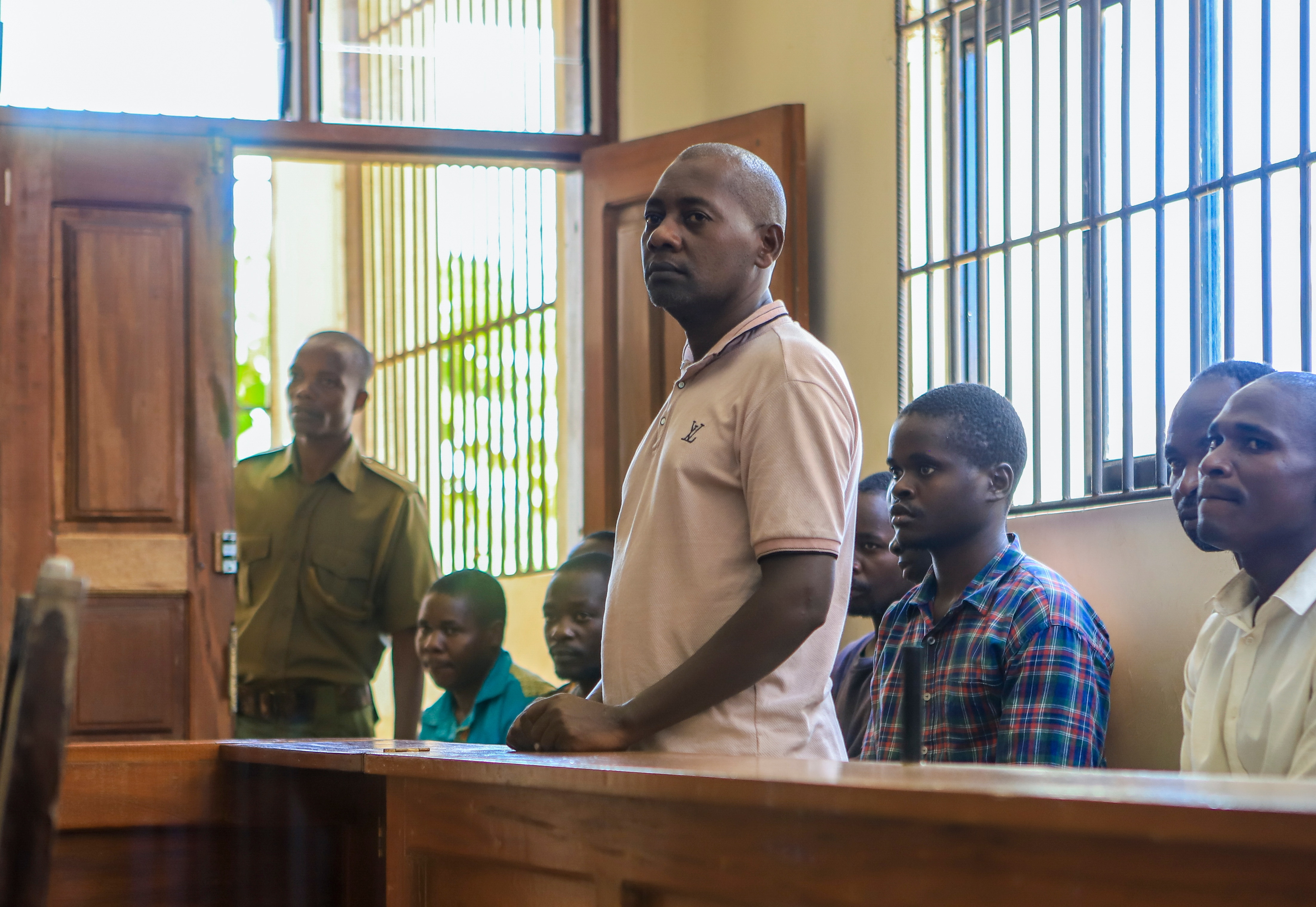 A middle-aged African man wearing a white-grey polo shirt stands behind a bench in a courtroom.
