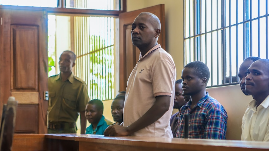 A middle-aged African man wearing a white-grey polo shirt stands behind a bench in a courtroom.