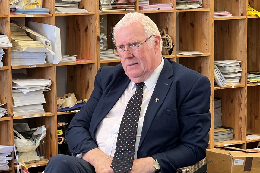 Eugene Reavey sits on a chair in a suit and tie in front of pigeon holes full of documents
