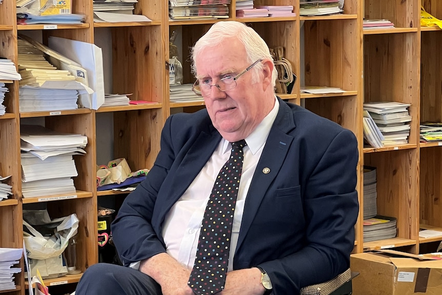 Eugene Reavey sits on a chair in a suit and tie in front of pigeon holes full of documents