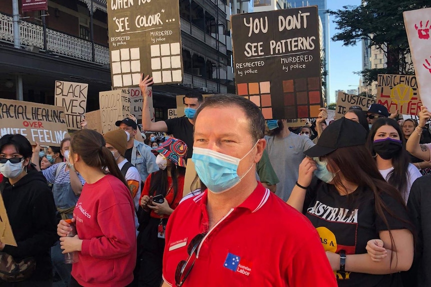 A man in a red shirt with Australian Labor on it wearing a mask in a crowd of people holding protest signs.