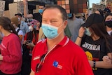 A man in a red shirt with Australian Labor on it wearing a mask in a crowd of people holding protest signs.
