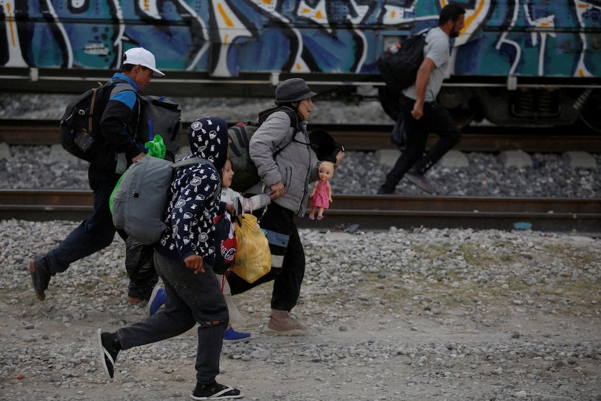 Five migrants, including a child, wearing backpacks and bags are pictured while running on a traintrack. 