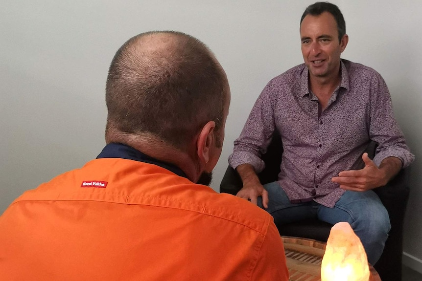 A man in an orange worker shirt, sitting with his back turned, speaking to a counsellor who is also seated