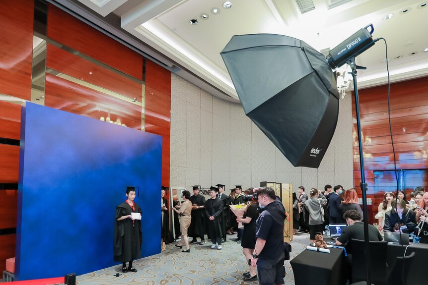 Students in black gowns line up to have their photo taken in a large room. 