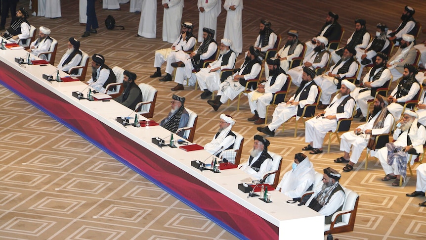 Men sit at one side of a long table in front of an audience of other men, some sitting and some in long robes standing nearby.