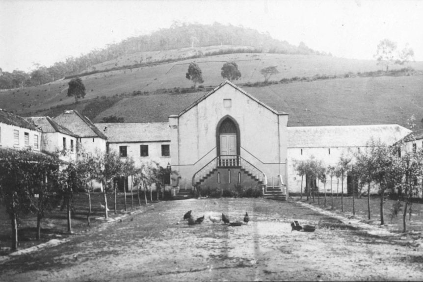 An historic black and white photo of a large sandstone building with a steep hill in the background and chickens in foreground.