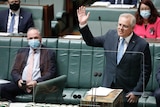 Barnaby Joyce sitting on a parliament bench, wearing a mask, while Scott Morrison stands with a hand in the air, speaking.