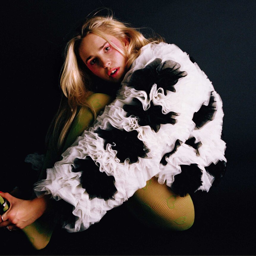 Laurel sits on a black background in a tulle black and white jacket holding her feet.