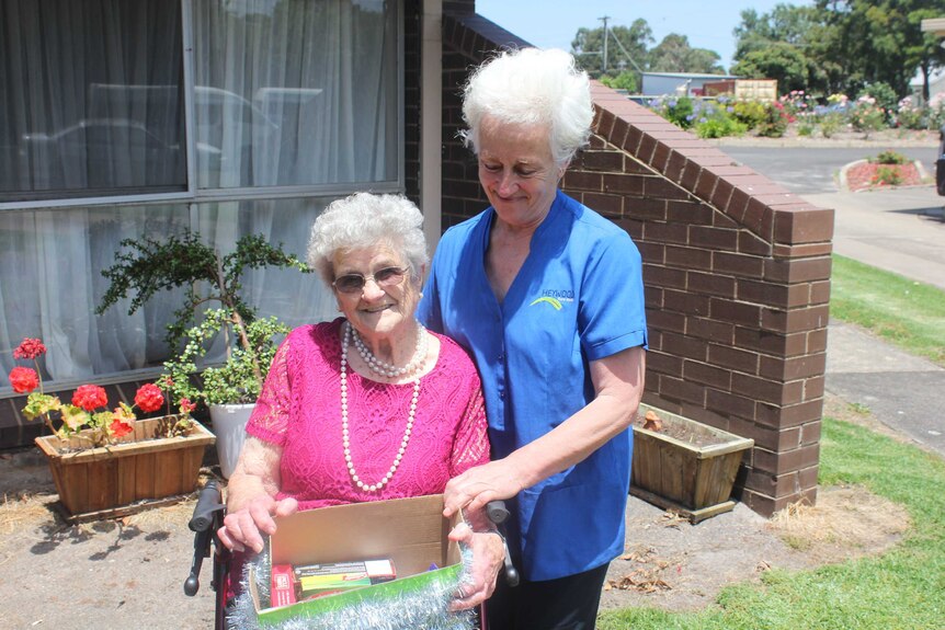 A nurse in a blue shirt stands next to an elderly lady sitting on a walker holding a box covered in tinsel.