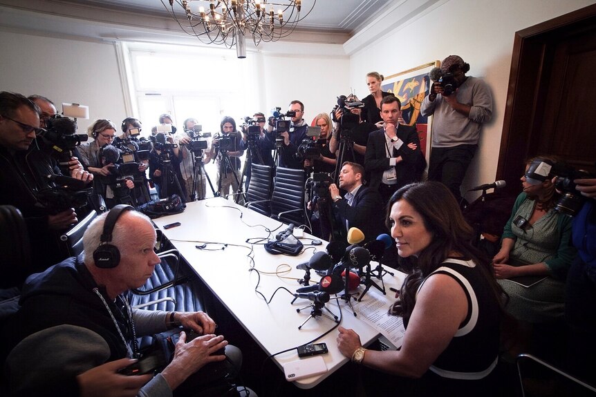 A woman sits at a table with several microphones, surrounded by reporters and camera operators.