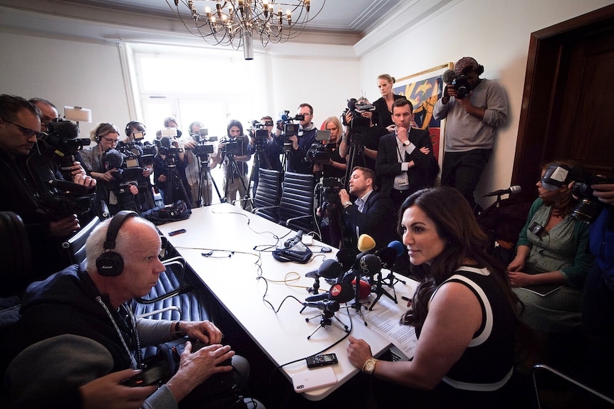 A woman sits at a table with several microphones, surrounded by reporters and camera operators.