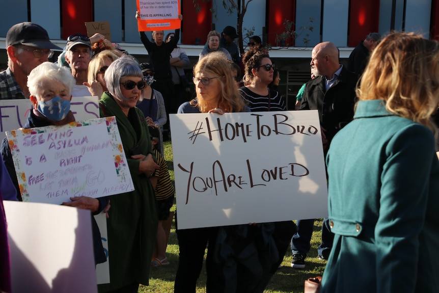 A woman in a crowd holds a placard that reads "Home to Bilo you are loved".