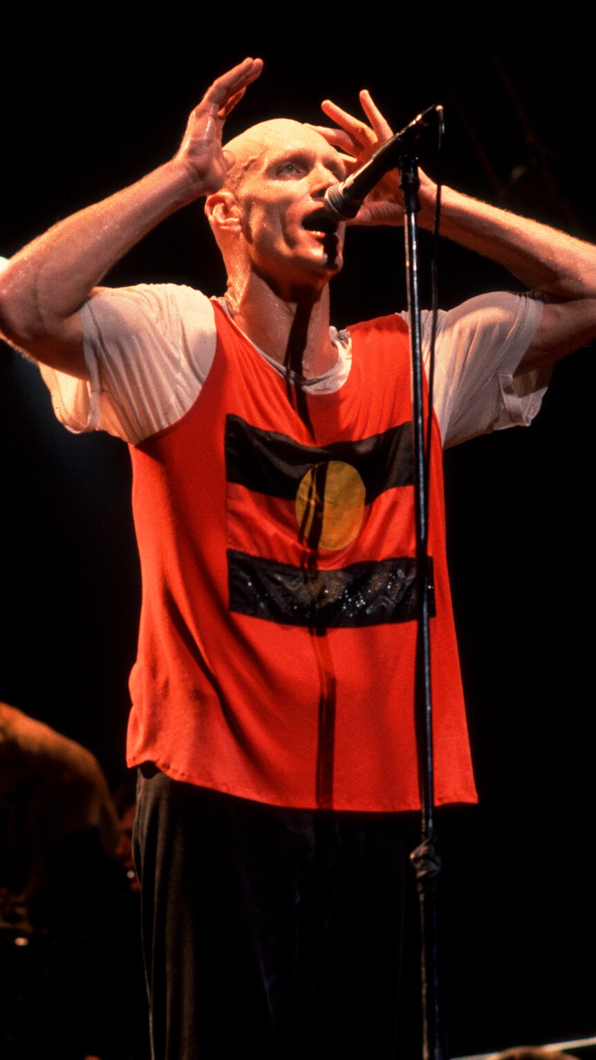 Peter Garrett sings into microphone. He is wearing black pants and a shirt that has an image of Aboriginal flag