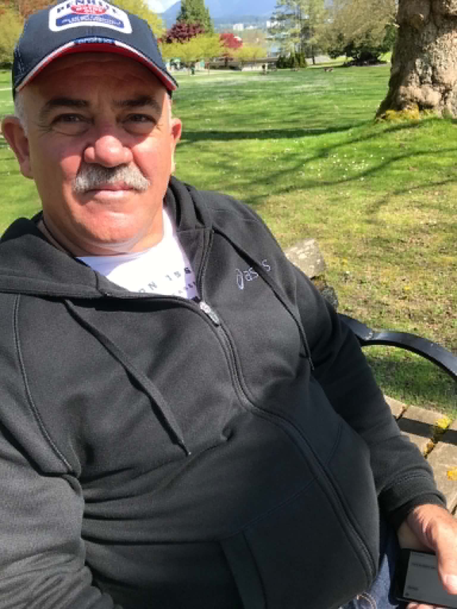 Older man with moustache, hat, jacket, on chair with green grass in background
