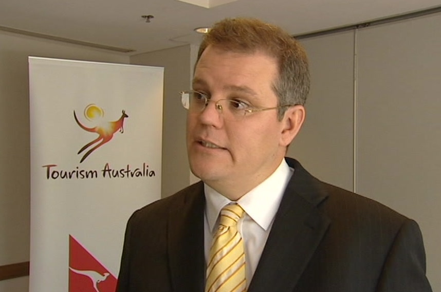 Scott Morrison giving a television interview with signage in the background showing logos of Tourism Australia and Qantas