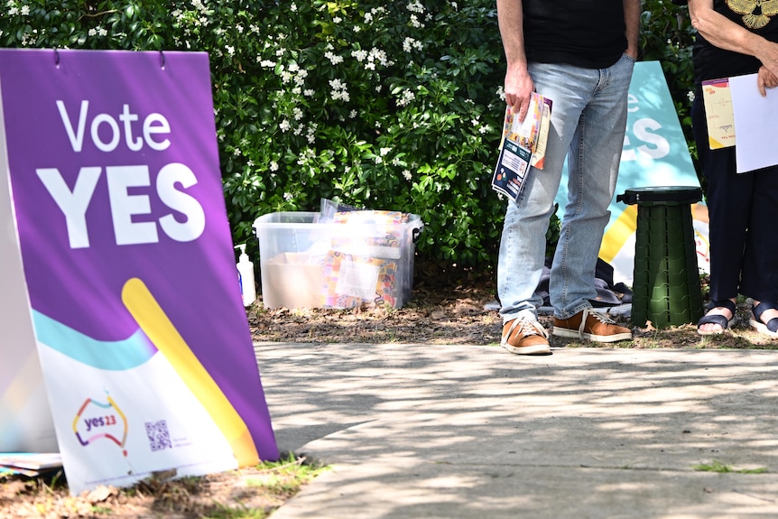 Three people stand in the background, between two purple and white YES signs