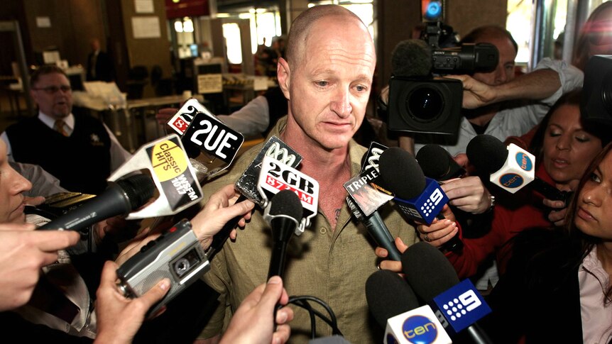 A man is surrounded by journalists with microphones and cameras.