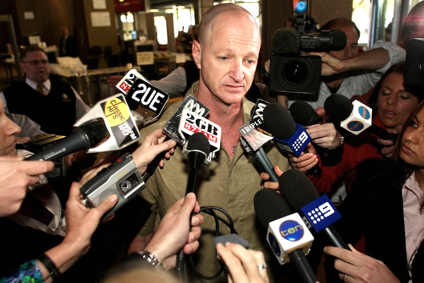 A man is surrounded by journalists with microphones and cameras.