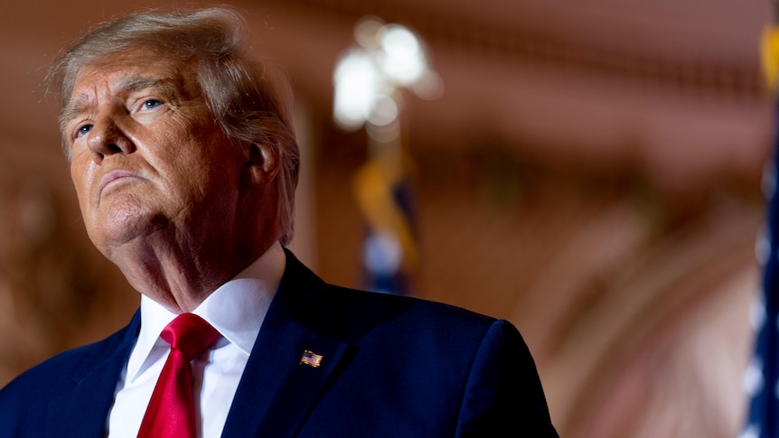 Donald Trump dressed in a red tie and suit stands in a ballroom.