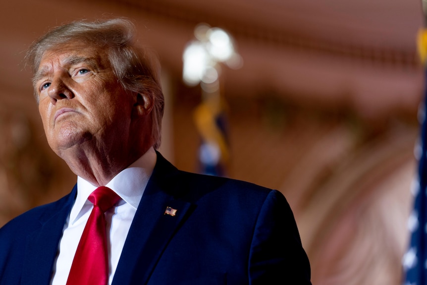 Donald Trump dressed in a red tie and suit stands in a ballroom.