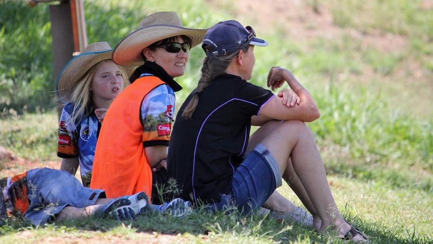 Two women in hats sit under a tree chatting a young girl in a cowboy hat watches on.