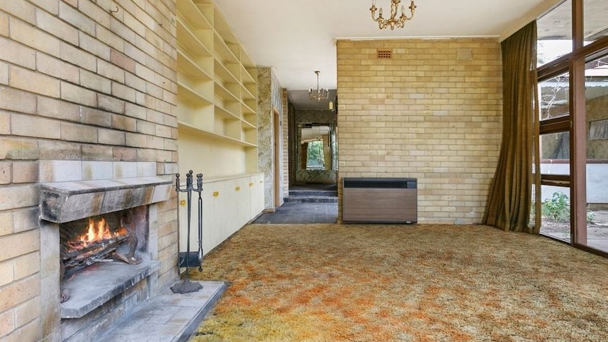 The living room of Gough Whitlam's former home has a fireplace and large bookshelf and floor-to-ceiling windows.
