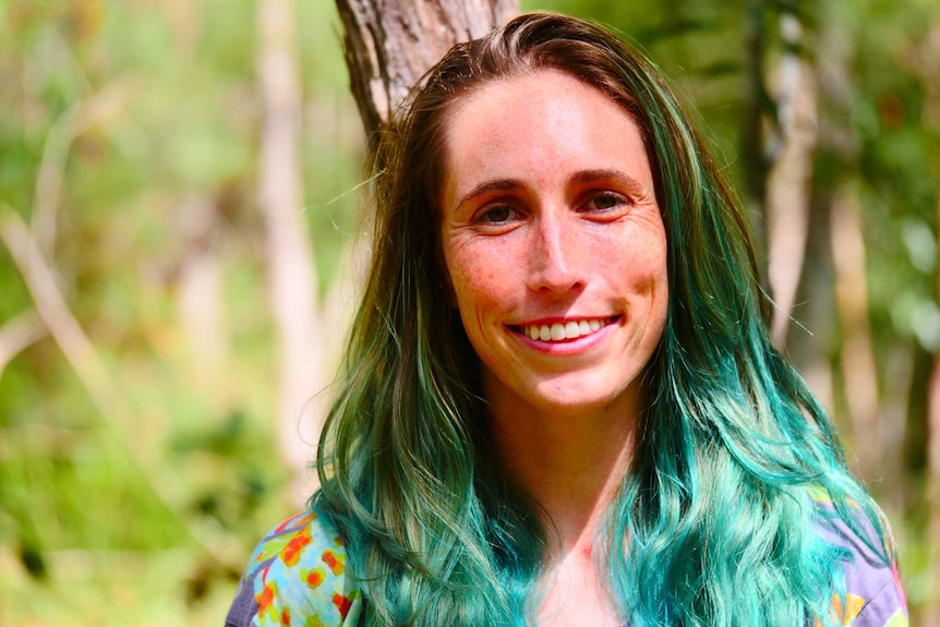 smiling woman with green dyed long hair and a bright top with trees behind her