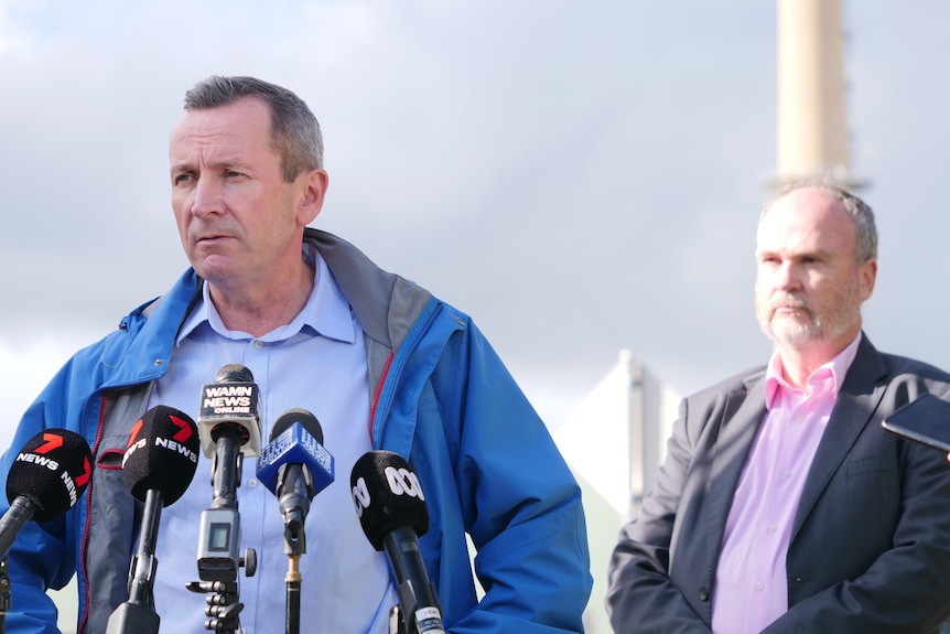 Mark McGowan speaks into microphones and Bill Johnston stands behind him in front of the power plant