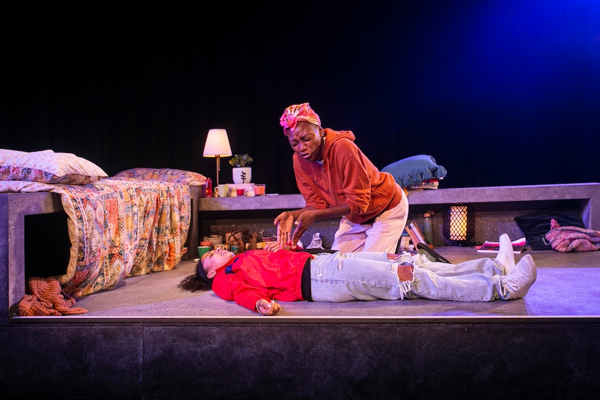One actor kneels beside another who is lying on her back in a room on a stage. The kneeling actor is gesticulating in distress.