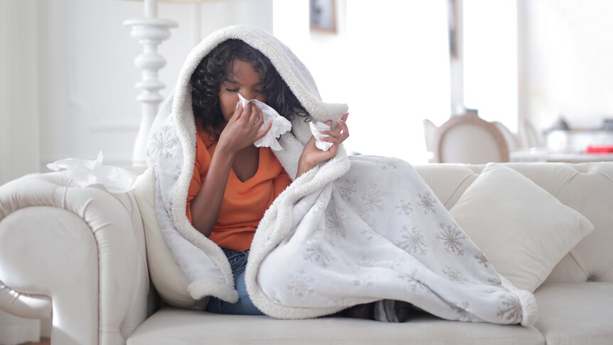 A woman sitting on a couch, wrapped in a cosy white blanket, sneezes into a tissue.