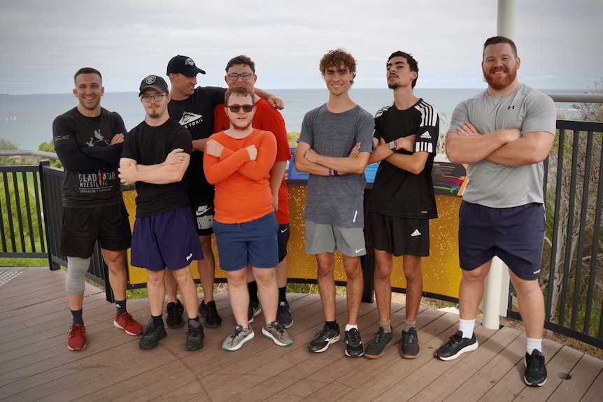 Nine men, dressed in fitness attire cross their arms and pose. They are on a deck outside. It is overcast.