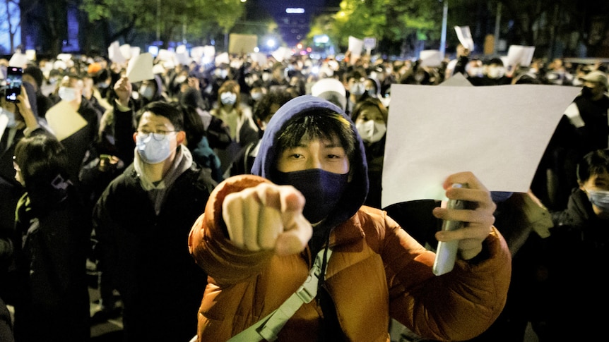 A crowd of people, some wearing masks, gather at night holding white sheets of paper in protest