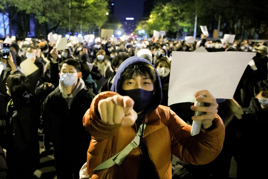 A crowd of people, some wearing masks, gather at night holding white sheets of paper in protest