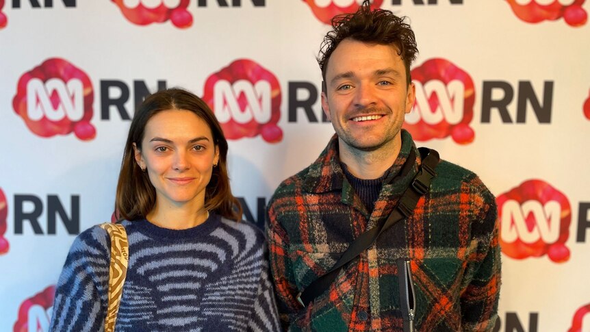 a women in a funky, printed jumper stands next to a man in a flannelette jacket . they smile at the camera