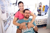 Ryan Marron sits smiling at the camera while his partner Toni Misitano stands behind him with her arms on his shoulders.