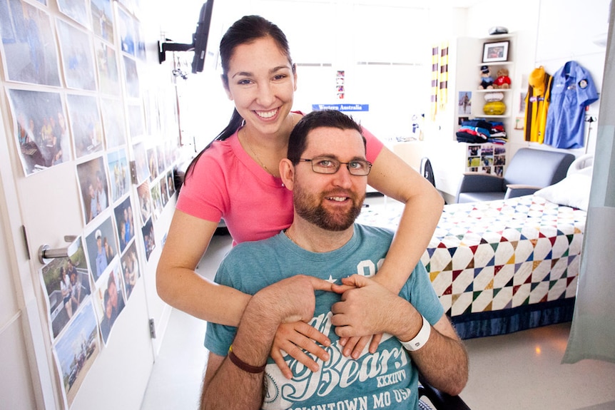 Ryan Marron sits smiling at the camera while his partner Toni Misitano stands behind him with her arms on his shoulders.