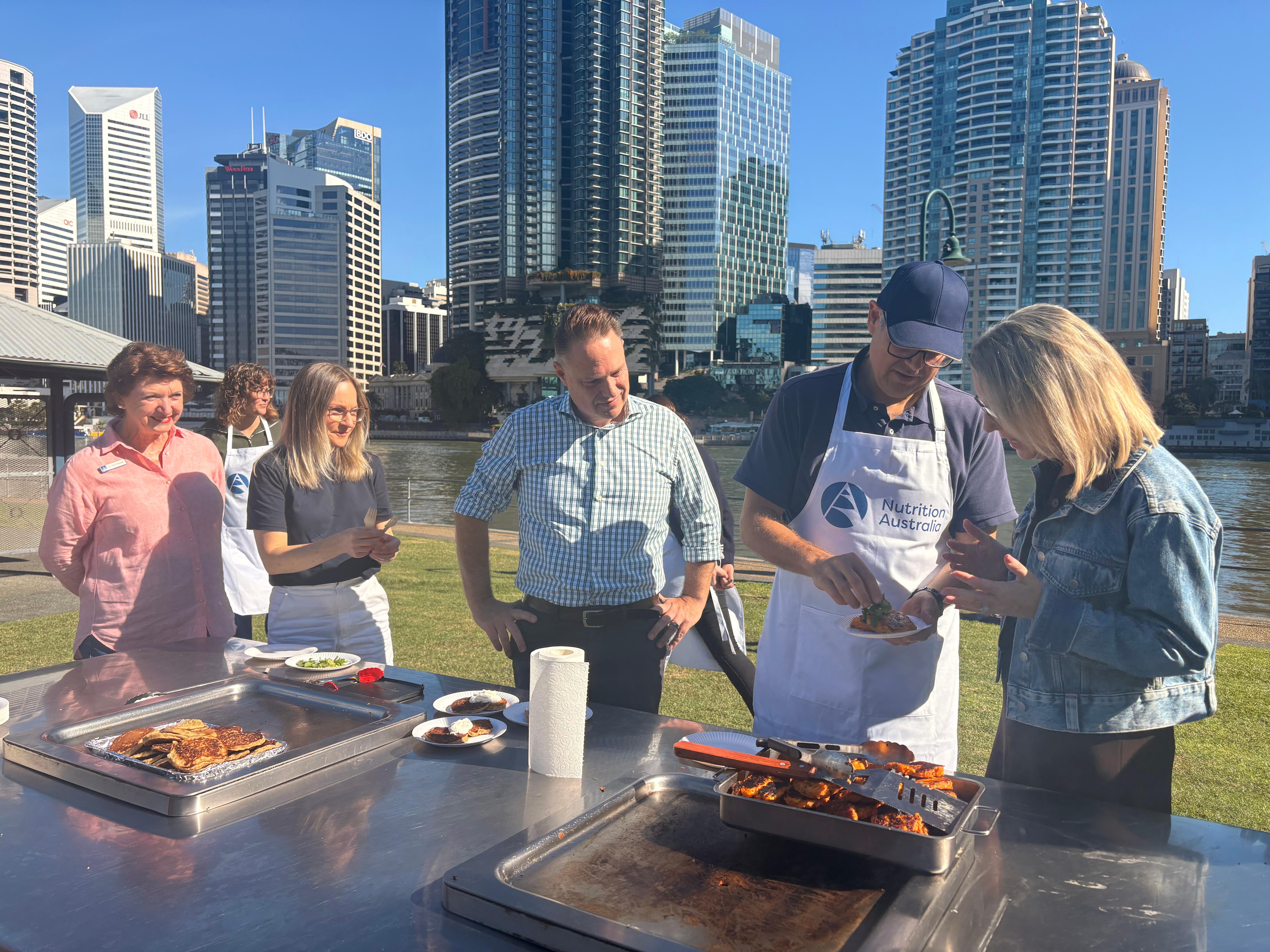 Three people, one in an apron, at a barbecue with Brisbane river and city in background.