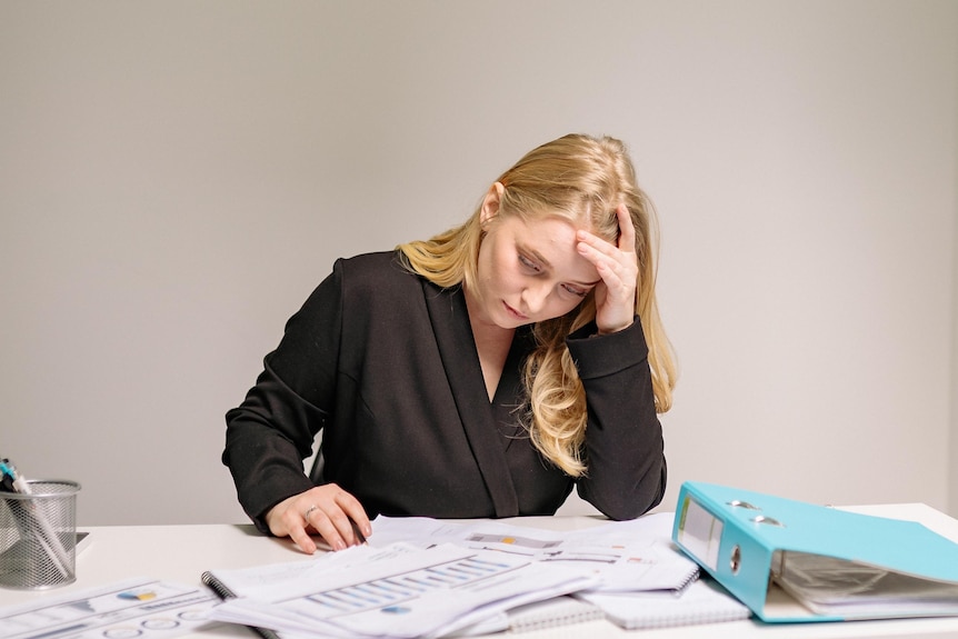A woman in a black top sits at a cluttered desk, looking stressed and tired