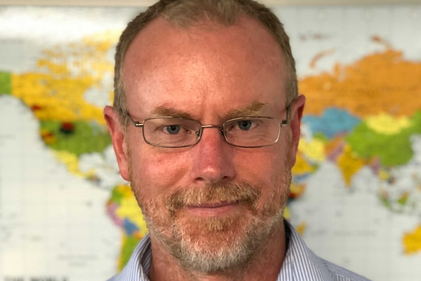 A head and shoulders image of a man with glasses and a beard standing in front of a world map.